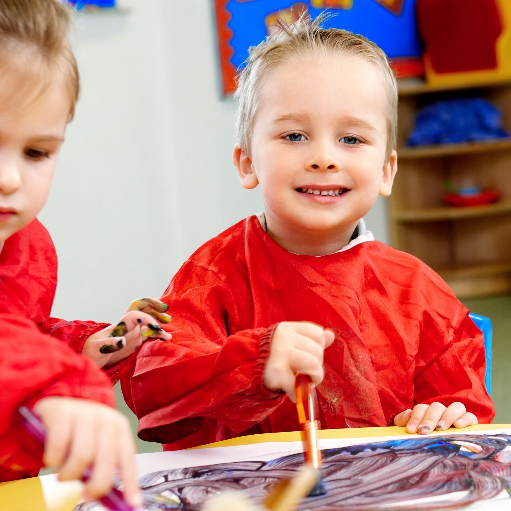 Smiling young boy painting at a table, wearing a red smock, inside a classroom setting.