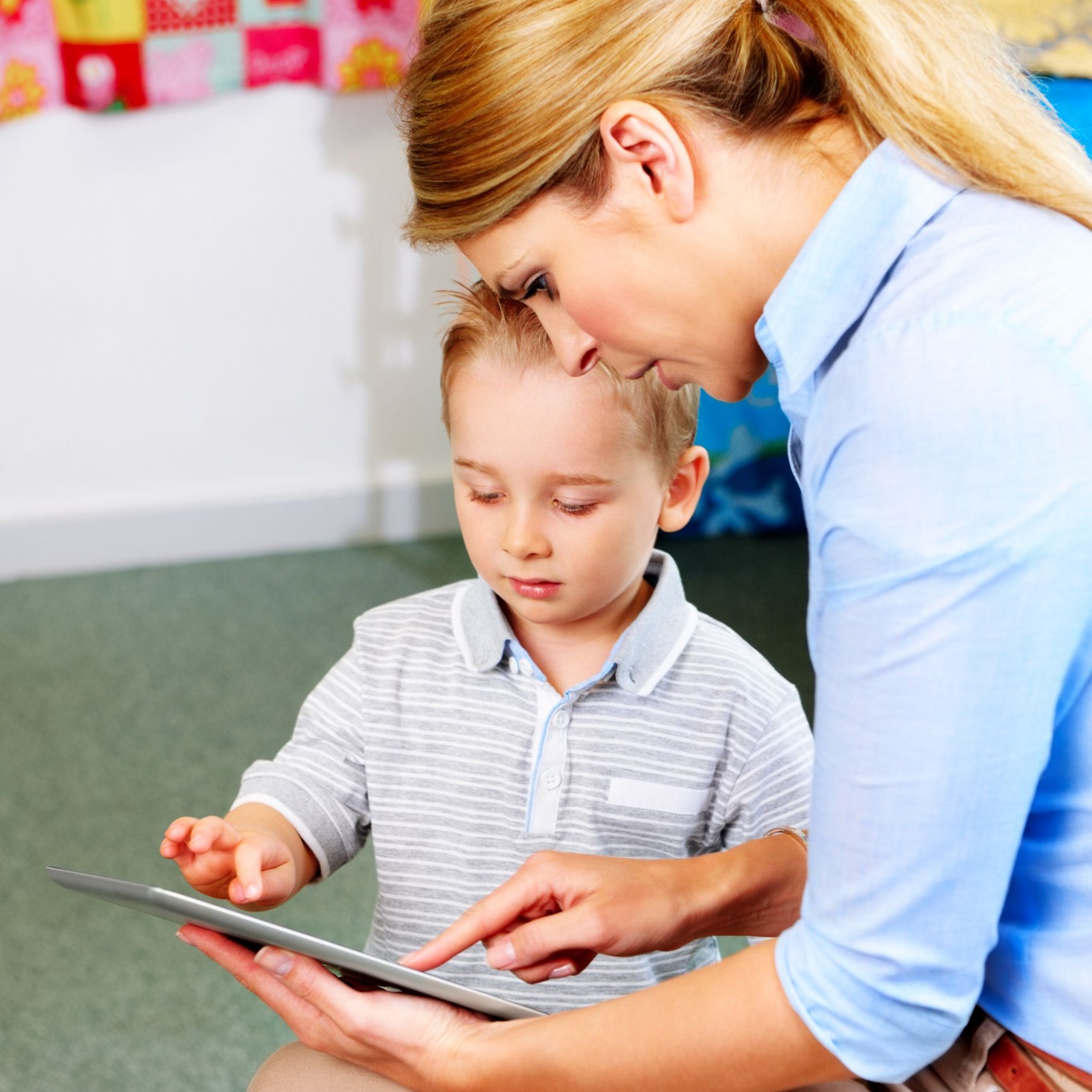 Woman in blue shirt helps a young boy in a grey shirt use a tablet indoors.