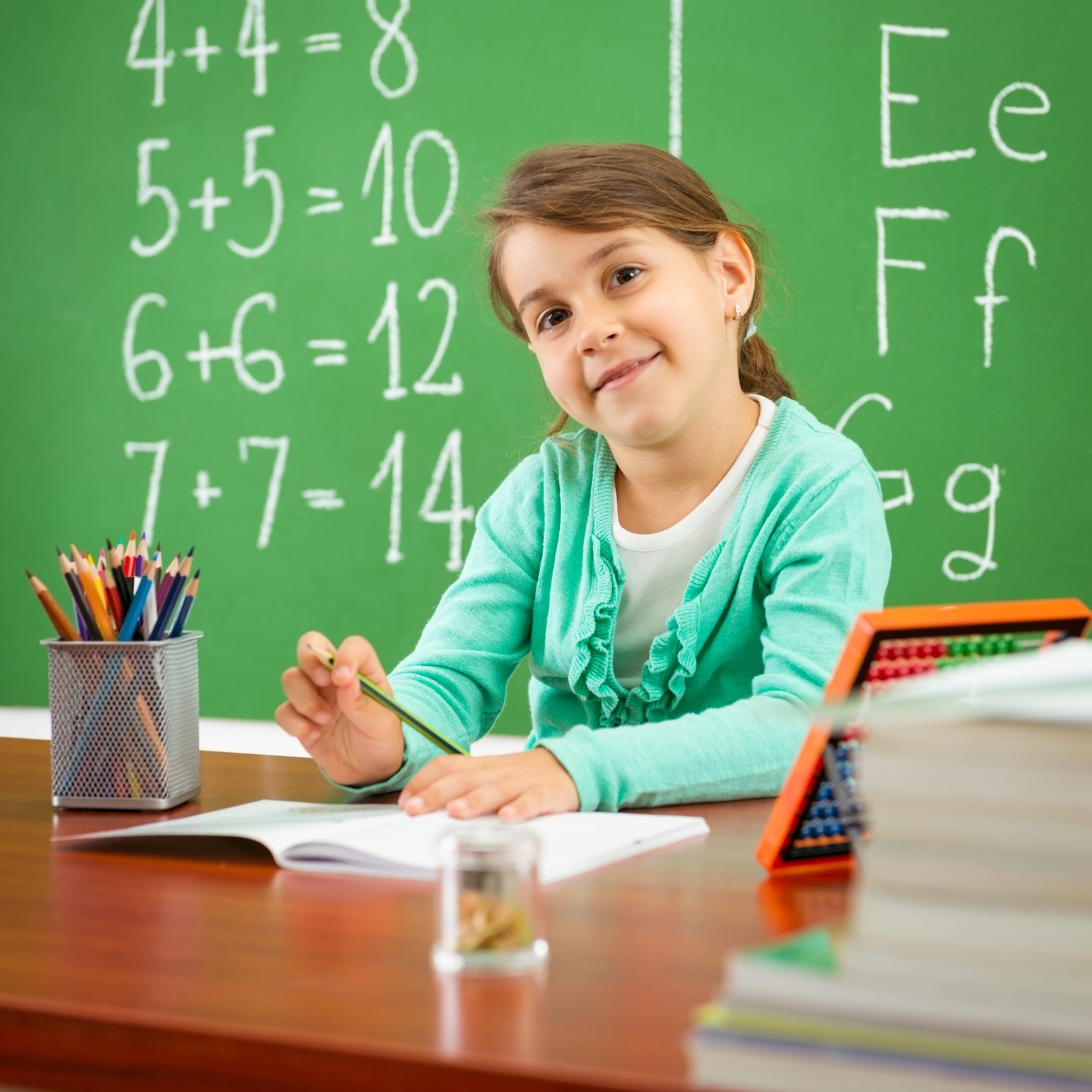 Girl in teal cardigan smiles, writing at desk in front of chalkboard with math problems.