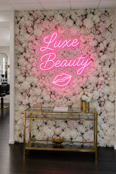 Pink neon “Luxe Beauty” sign and lips on a white floral wall above a gold console table.