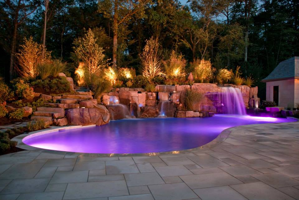 Nighttime view of a pool with purple lighting, waterfalls, and illuminated landscaping.
