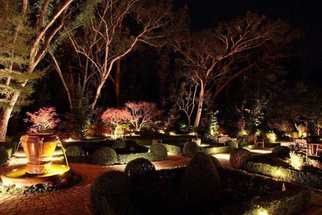 A landscaped garden at night, illuminated by warm yellow lights, featuring a stone fountain and manicured hedges.