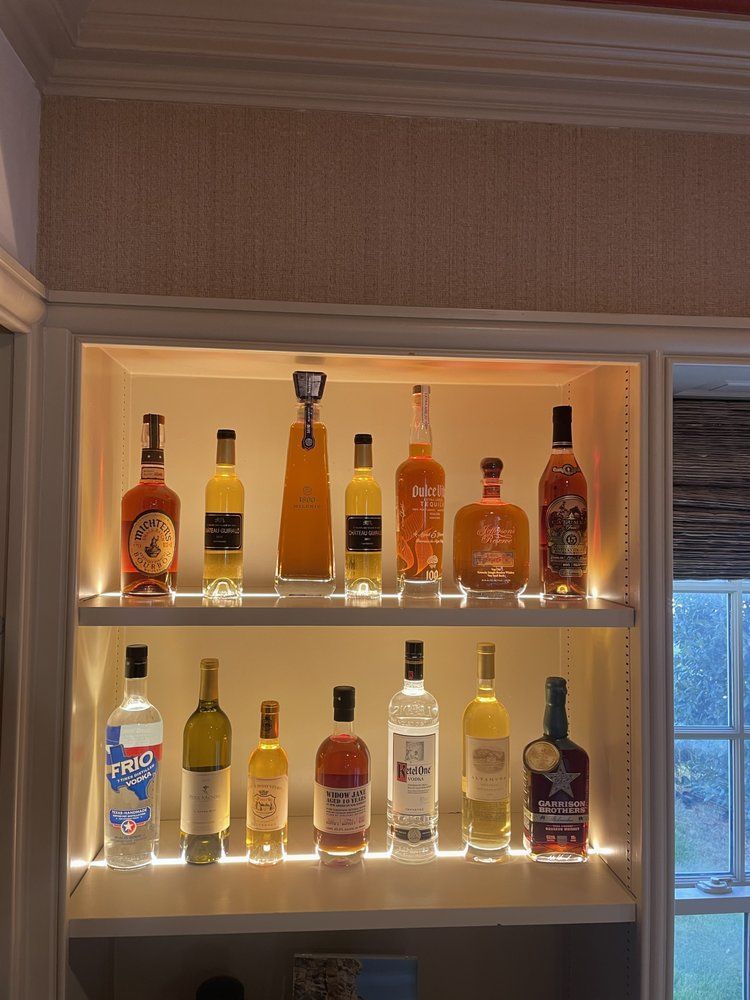 Two illuminated shelves displaying a collection of various liquor and wine bottles in a home bar setting.