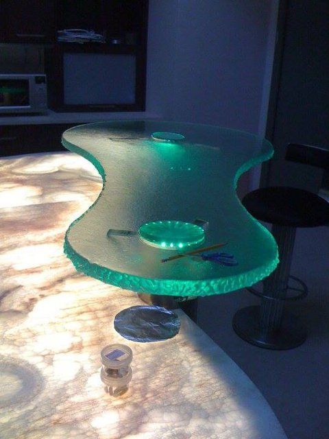 A translucent, glowing green glass table with textured edges sits atop a backlit, marble-patterned surface in a dark room.