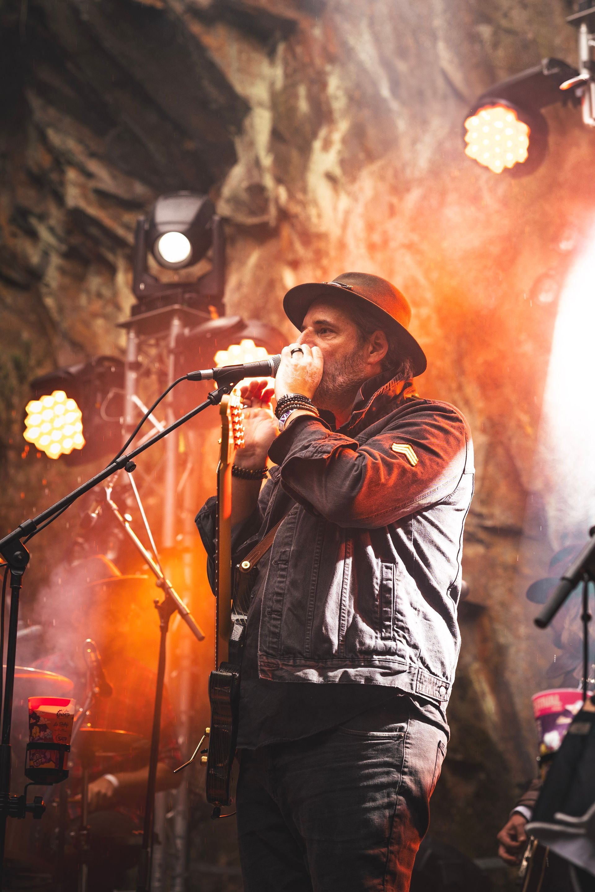 Un musicien coiffé d'un chapeau joue de l'harmonica et du micro sur une scène à l'éclairage chaleureux et tamisé, dans une ambiance rock.