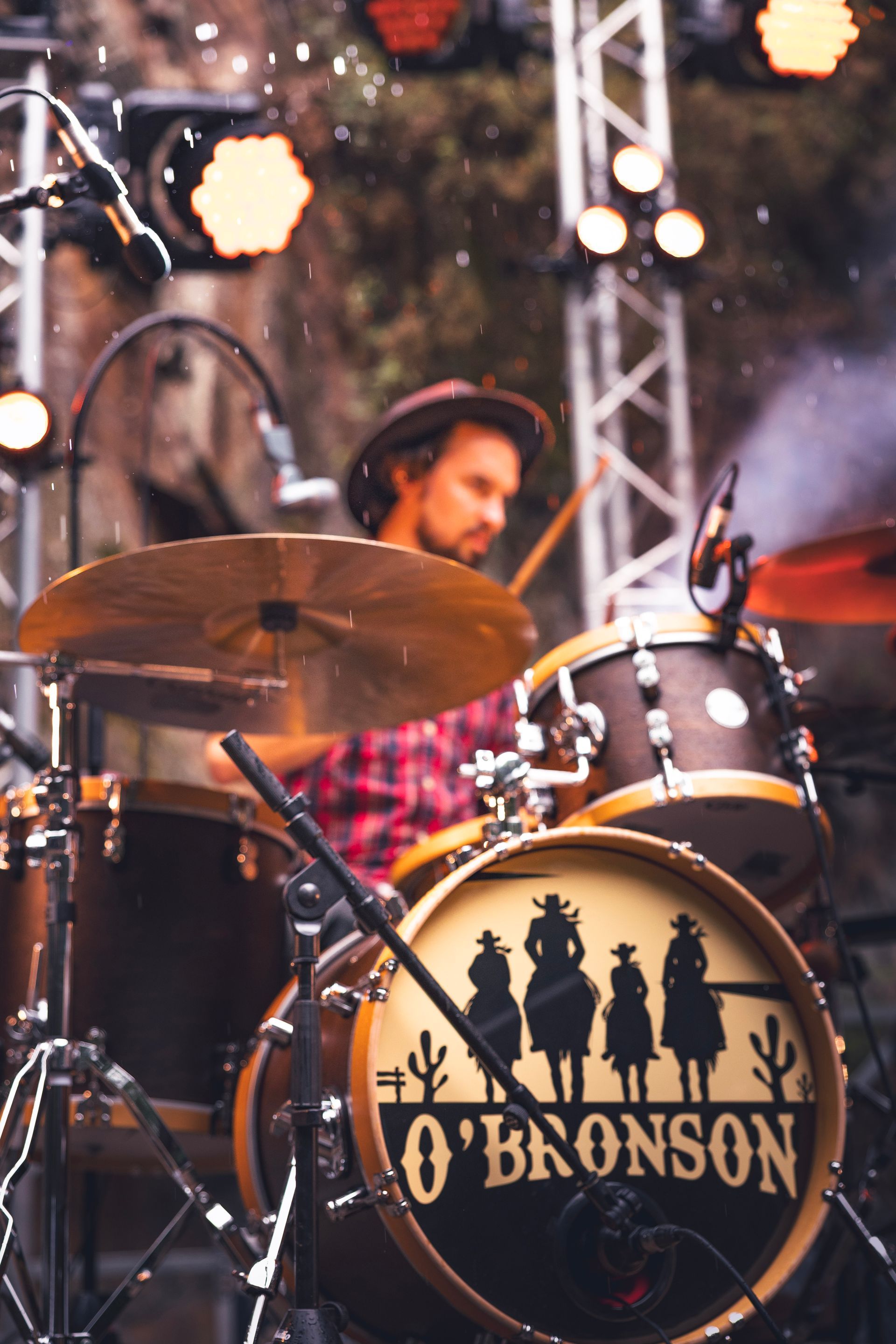 Un batteur joue sur une batterie marquée « O'BRONSON » ornée d'une silhouette de cow-boy, sur fond de scène éclairée par des projecteurs puissants.