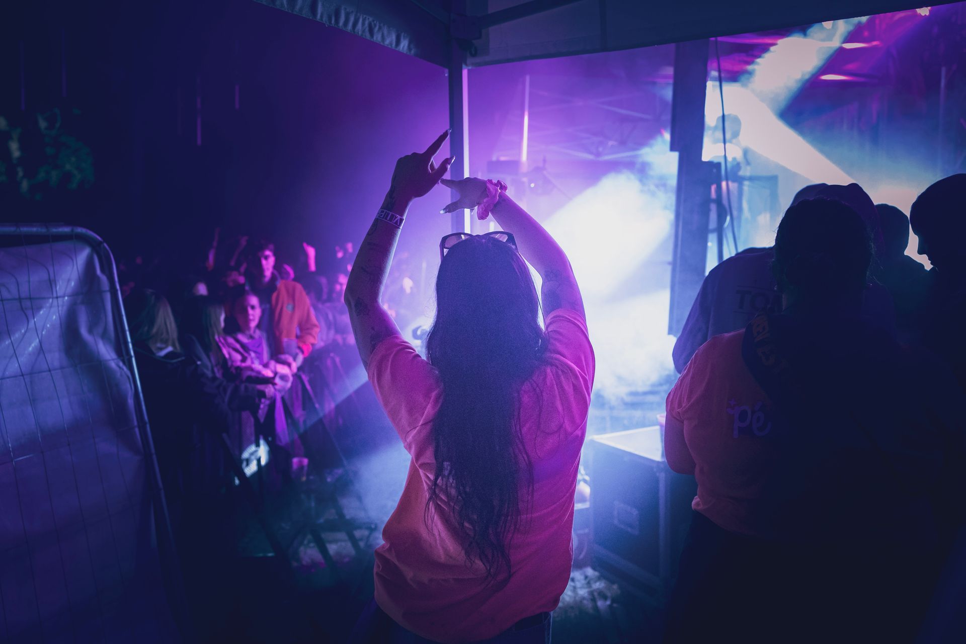 Une personne vêtue d'un t-shirt rose vif lève les bras lors d'un événement musical où règnent un éclairage de scène violet éclatant et de la fumée.