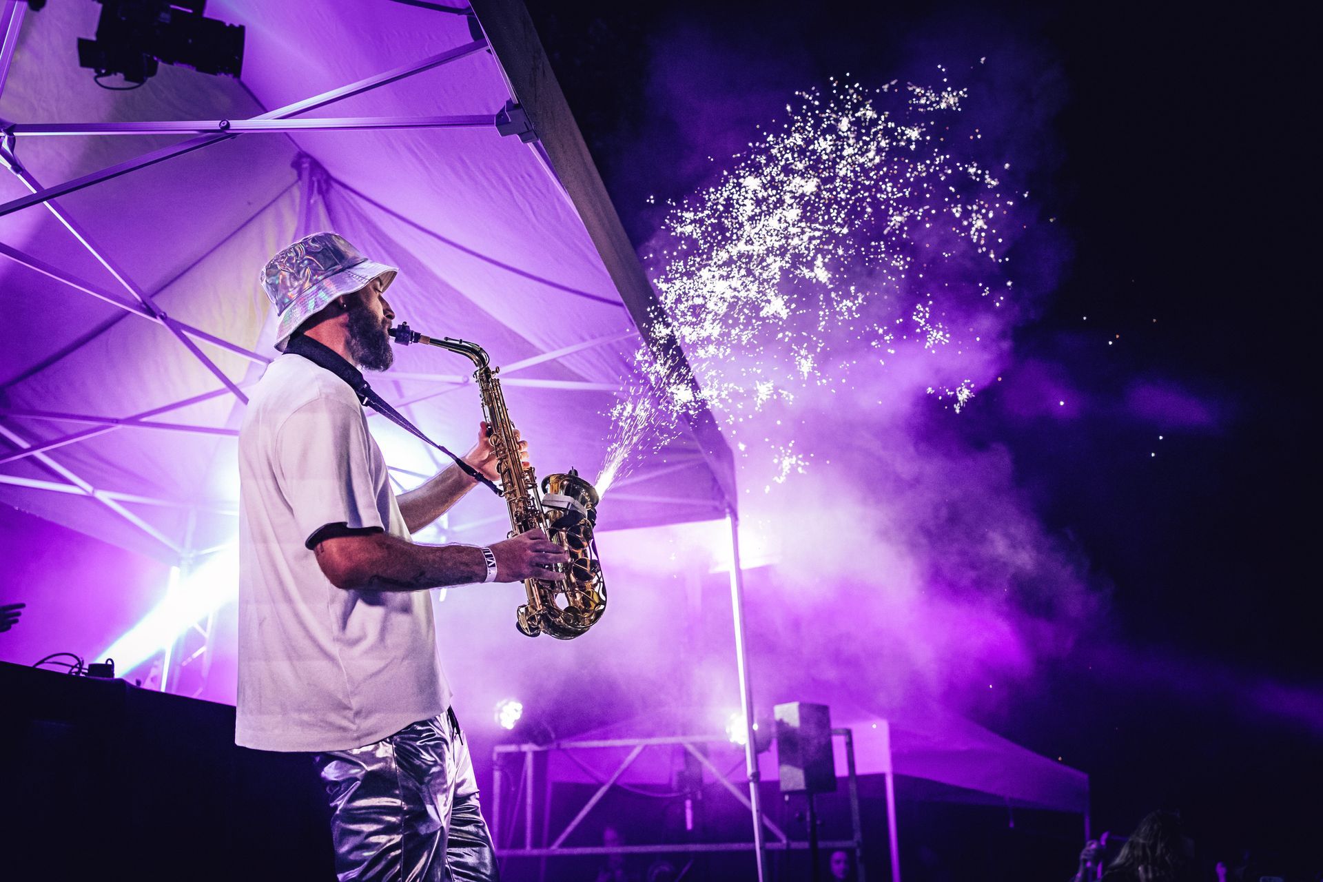 Un musicien coiffé d'un bob à motifs joue du saxophone sur scène, sous des lumières violettes, au milieu d'effets pyrotechniques scintillants.