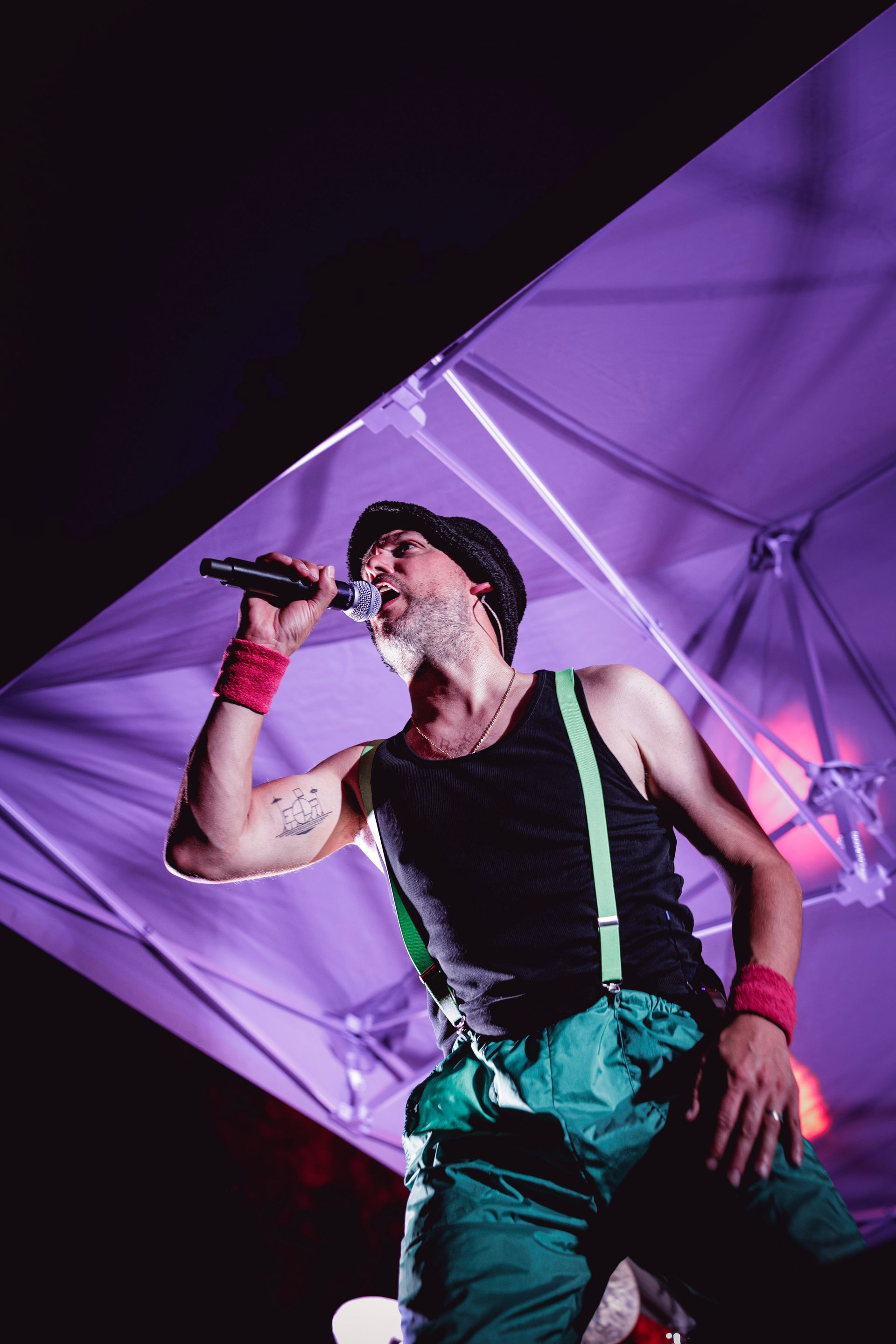 Un artiste chante dans un micro sur scène, vêtu d'un débardeur noir, d'un pantalon vert, de bretelles et de bracelets.