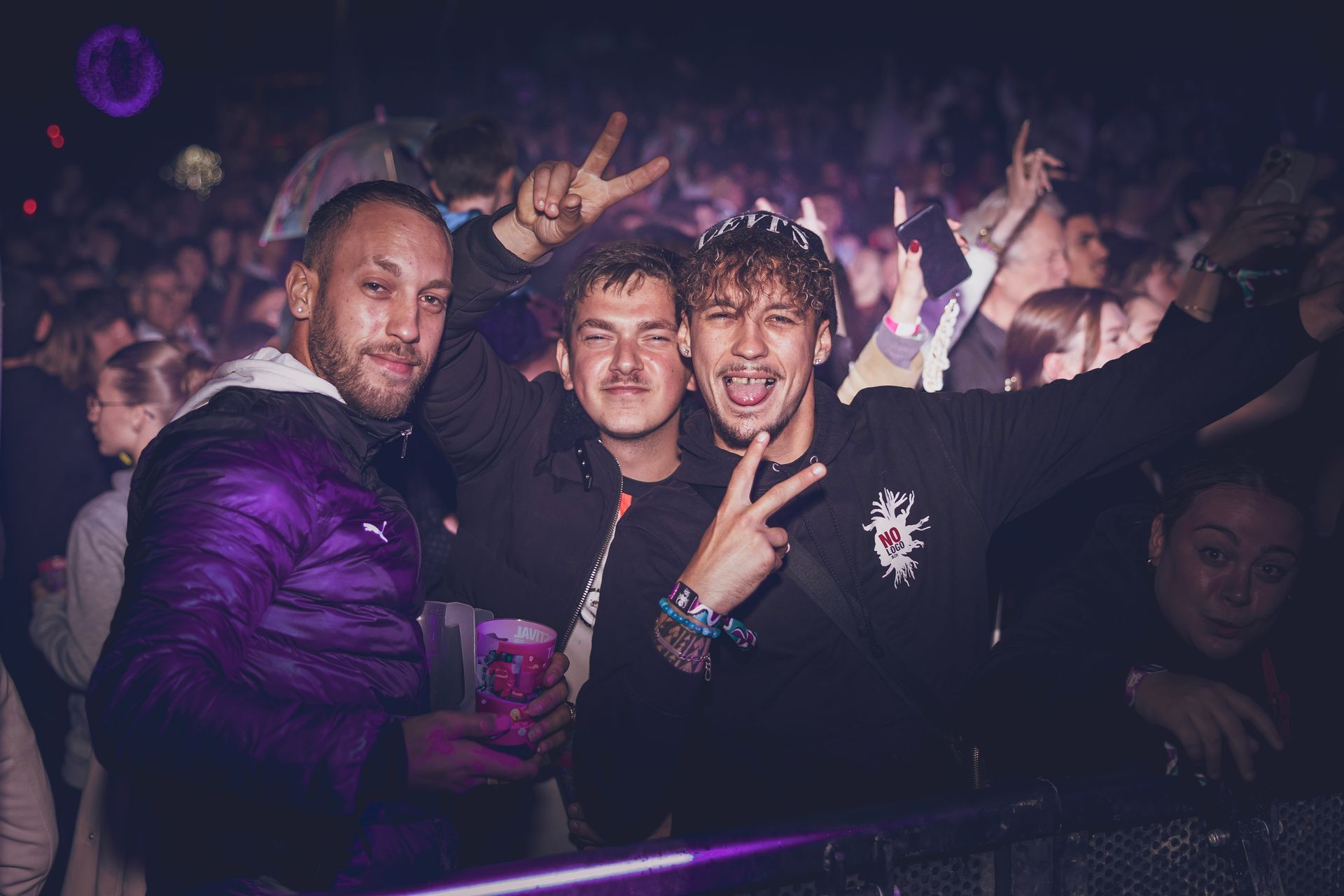 Trois personnes posent pour une photo dans une boîte de nuit bondée et faiblement éclairée, deux d'entre elles faisant le signe de la paix et tenant un verre.