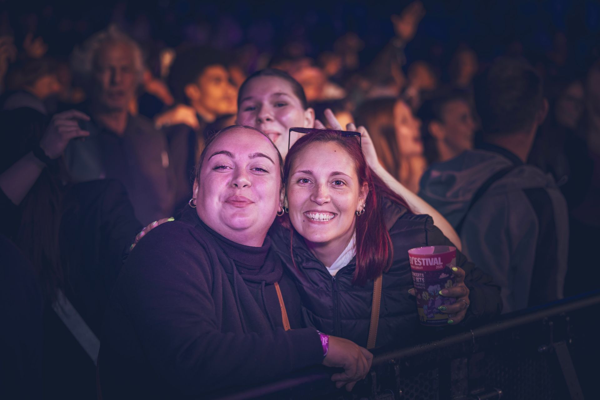 Dans une salle de concert bondée et faiblement éclairée, trois personnes sourient étroitement ensemble, l'une d'elles tenant un verre.