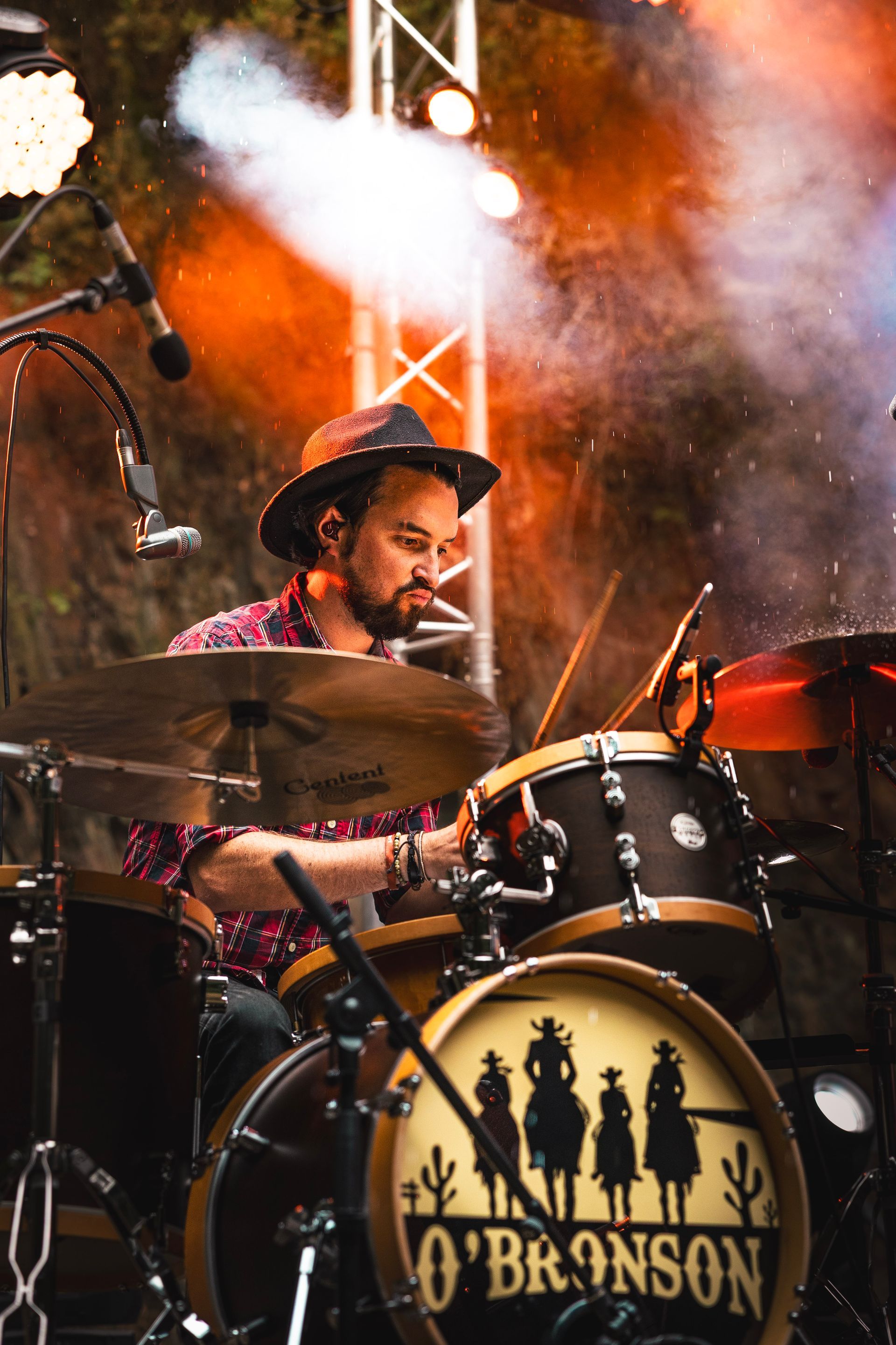 Un batteur coiffé d'un chapeau joue sur une batterie ornée du logo « O'Bronson » sur la grosse caisse, éclairée par des projecteurs et de la fumée.