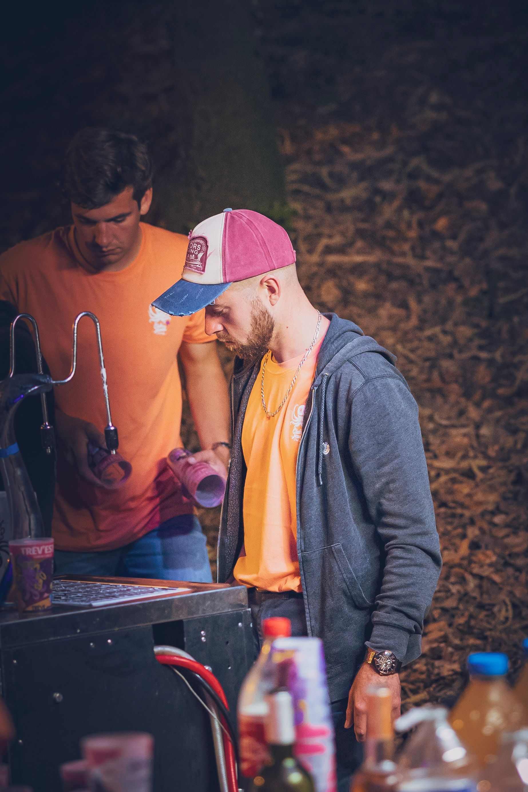 Deux personnes vêtues de chemises orange travaillent derrière un bar la nuit, l'une d'elles portant une casquette de baseball rose et bleue.