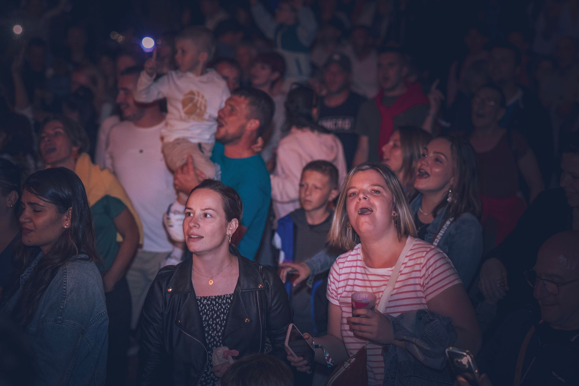 Dans une ambiance tamisée, une foule hétéroclite assiste à un événement, les yeux rivés sur l'événement, l'air concentré et enthousiaste.