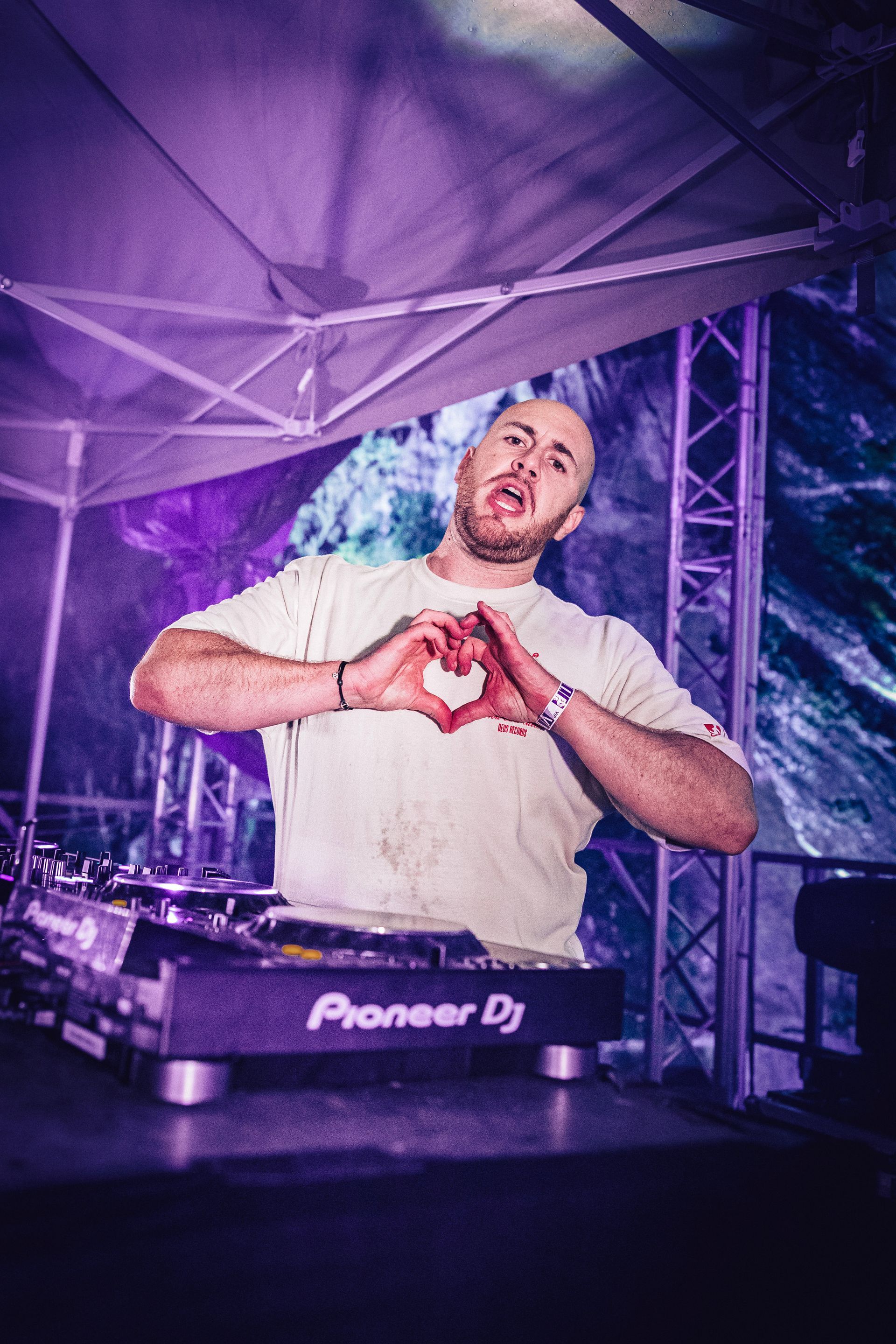 Un DJ derrière une console Pioneer DJ lors d'un concert, formant un cœur avec ses mains et regardant le public.