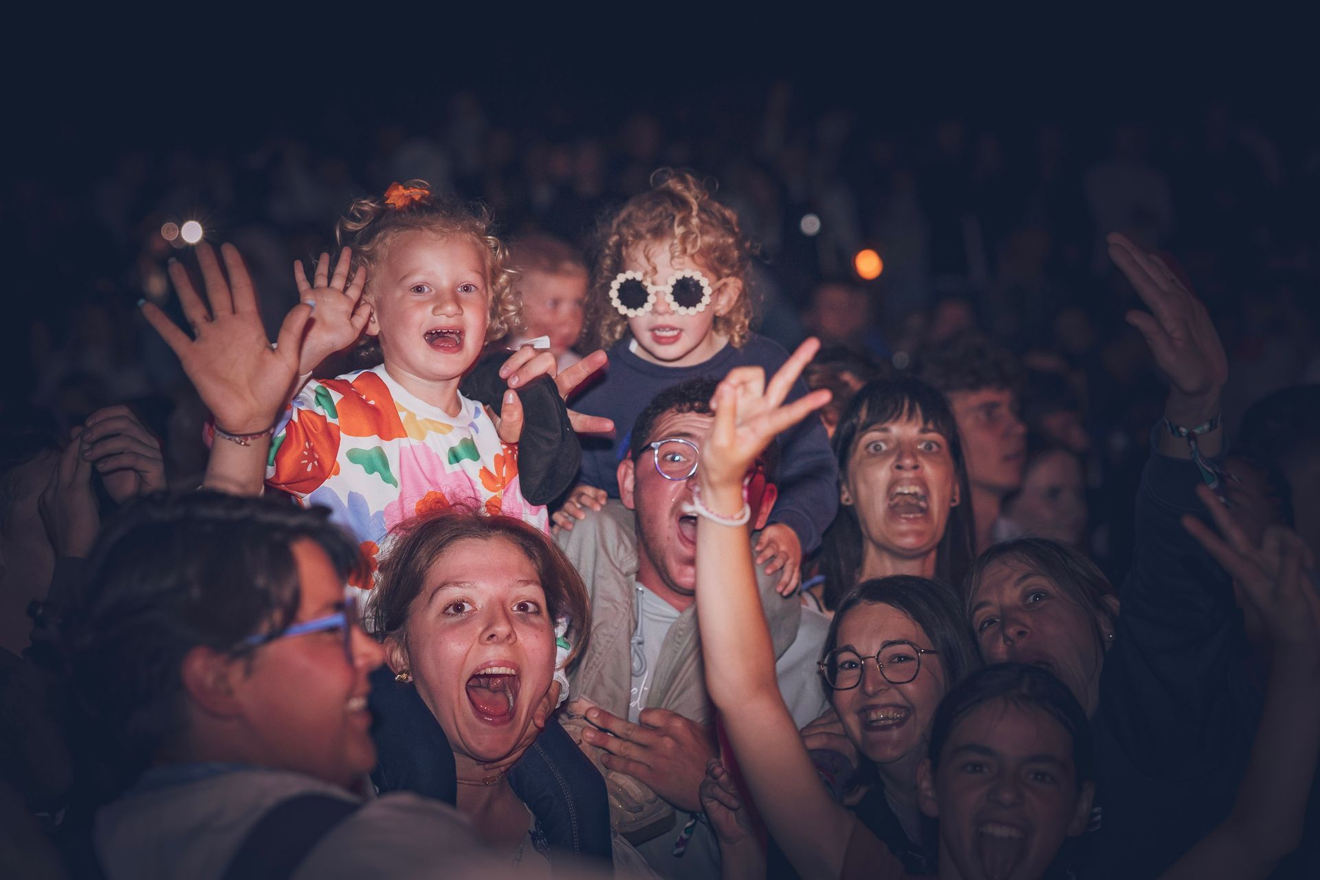 Une foule hétéroclite lors d'un concert en soirée : les gens applaudissent, sourient et font des signes de la main, certains portant des lunettes de soleil.
