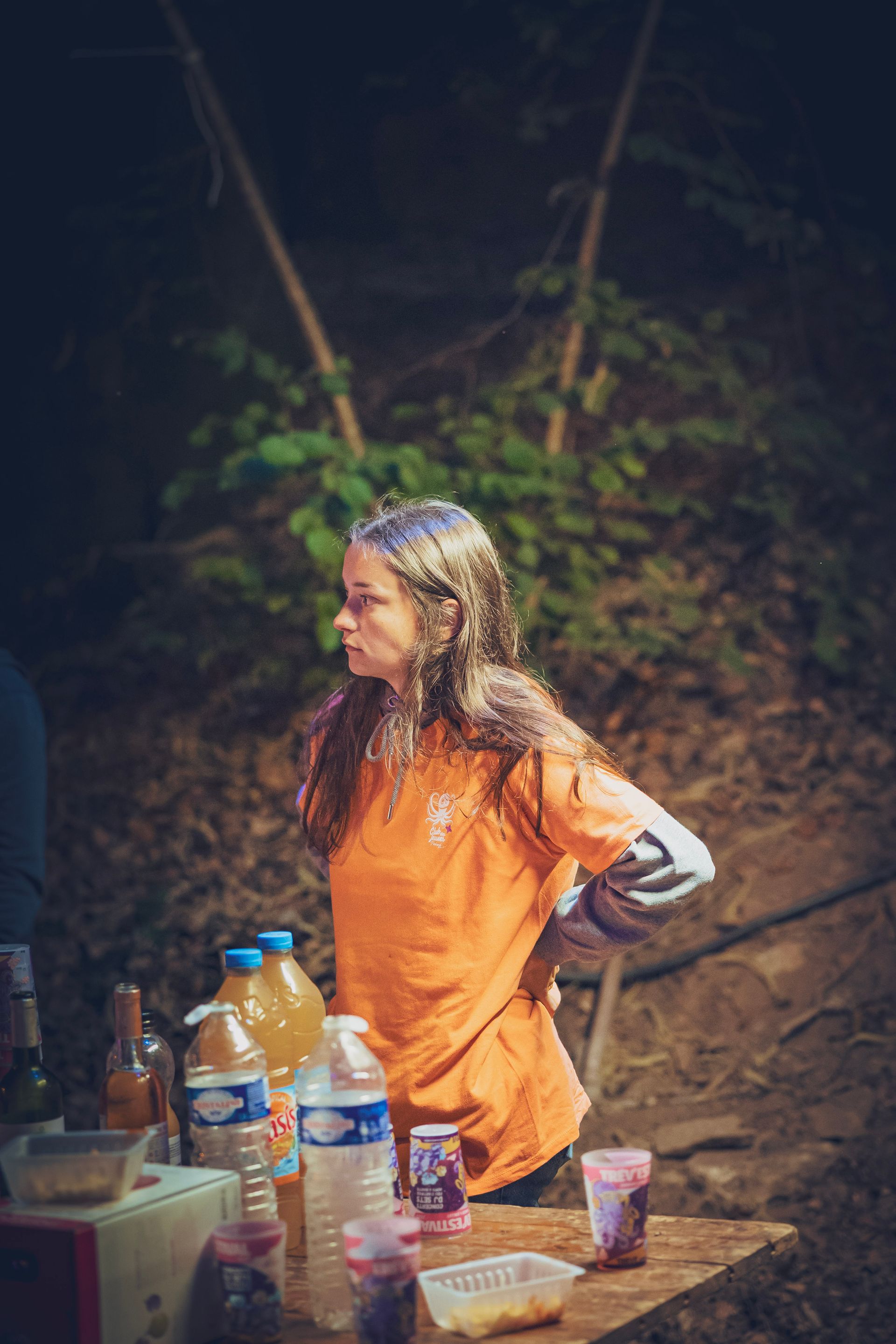 Une personne vêtue d'un t-shirt orange se tient derrière une table avec des bouteilles et des verres, dans un décor boisé de nuit.