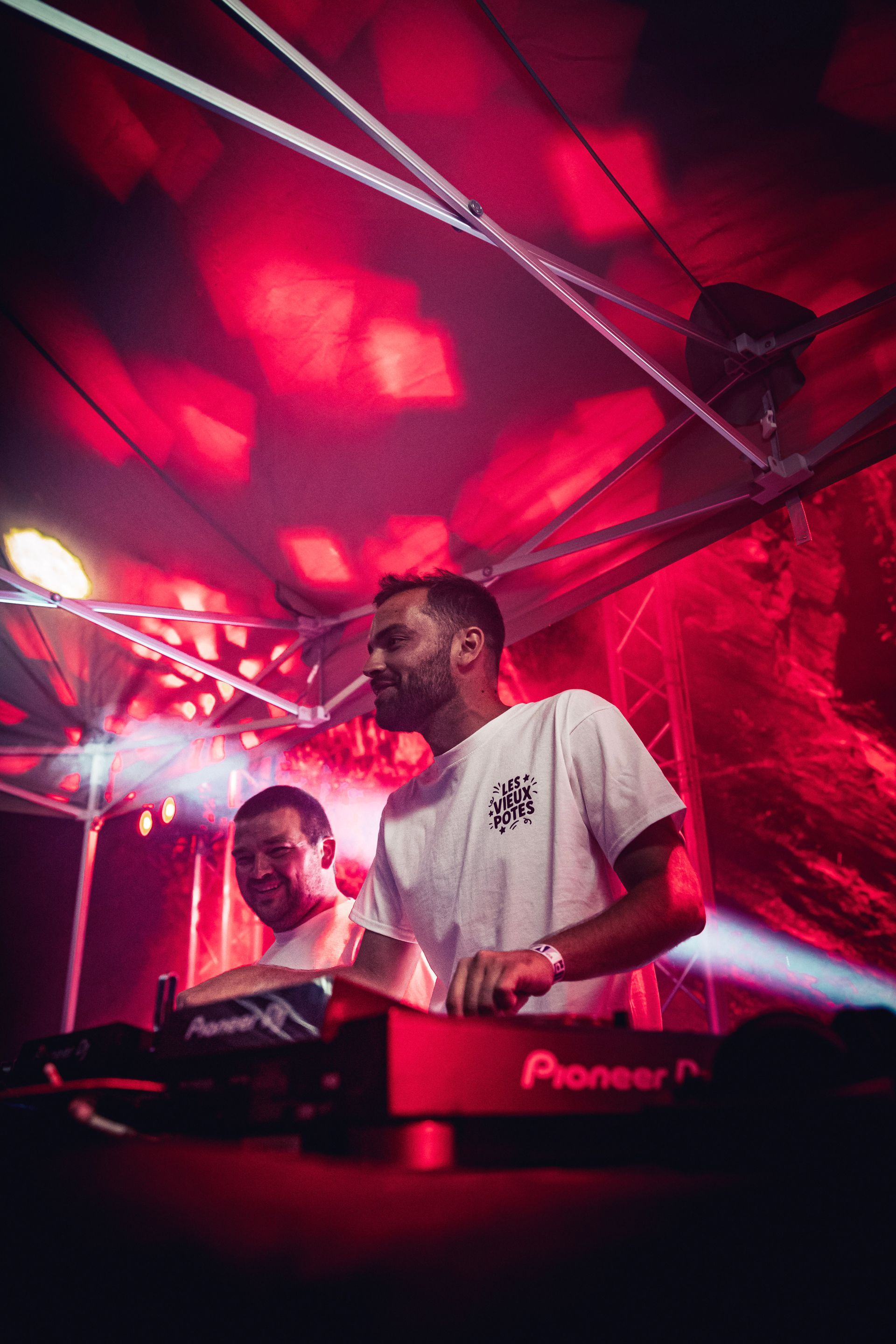 Deux personnes manipulent du matériel de DJ dans une salle de concert faiblement éclairée aux teintes rouges, avec un éclairage professionnel.