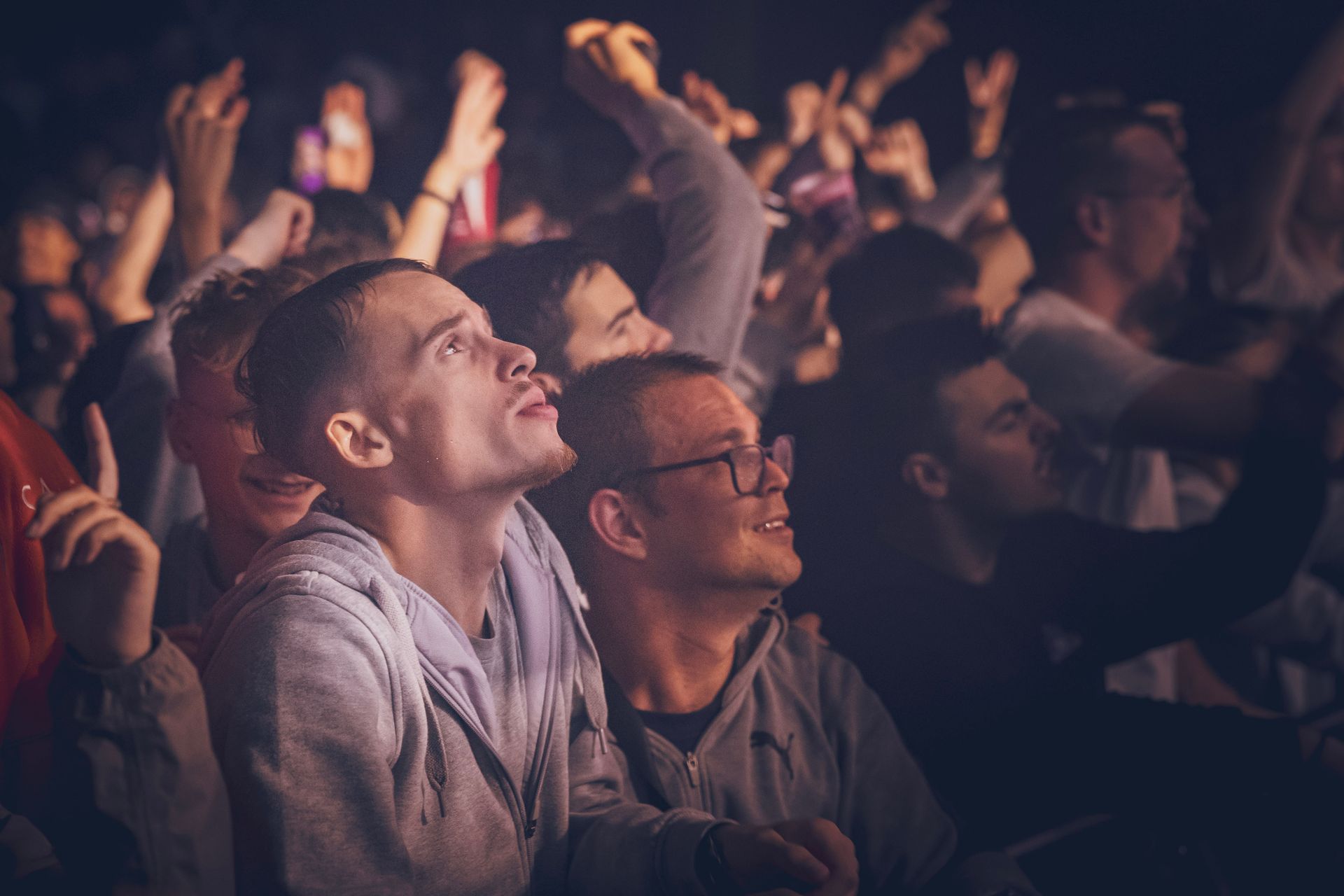 Une foule assiste à un spectacle dans une salle obscure, certains individus levant les yeux au ciel avec des expressions concentrées.