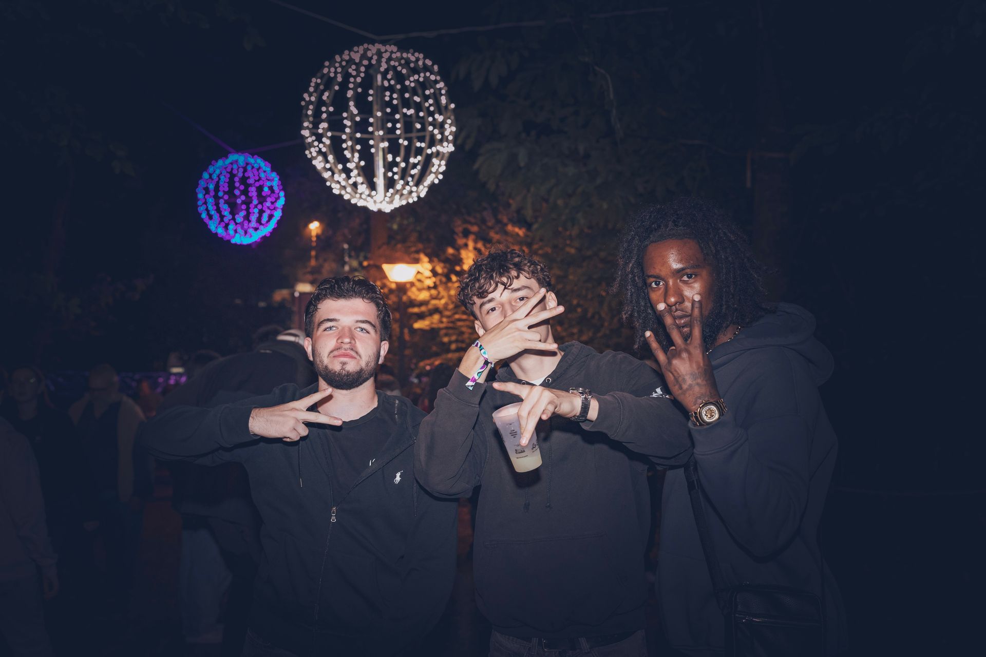 Trois personnes vêtues de sweats à capuche sombres posent pour une photo de nuit, éclairées par des lumières sphériques scintillantes en arrière-plan.