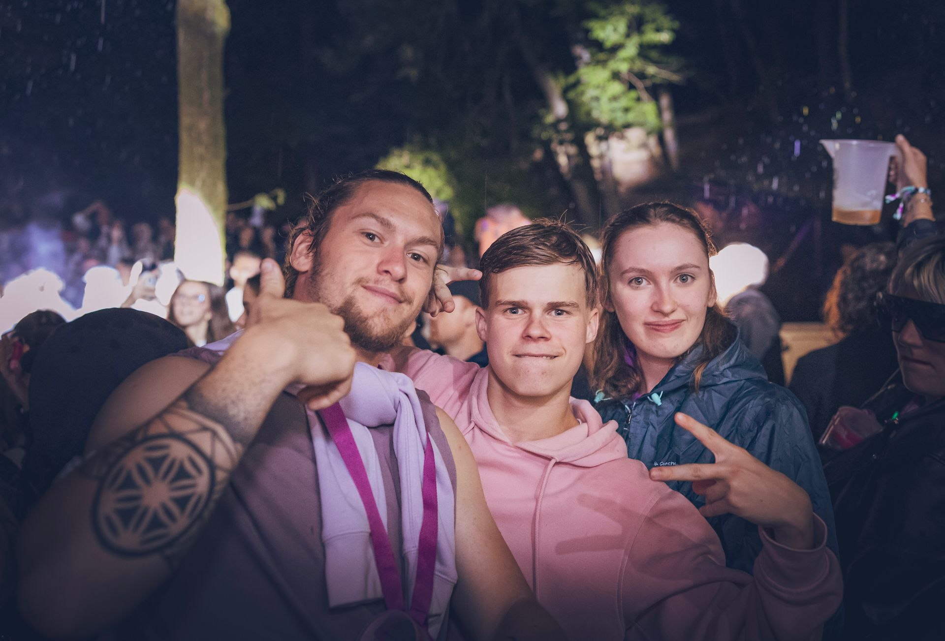 Trois personnes posent ensemble lors d'un événement nocturne en extérieur, celle de gauche faisant un geste et montrant un tatouage.