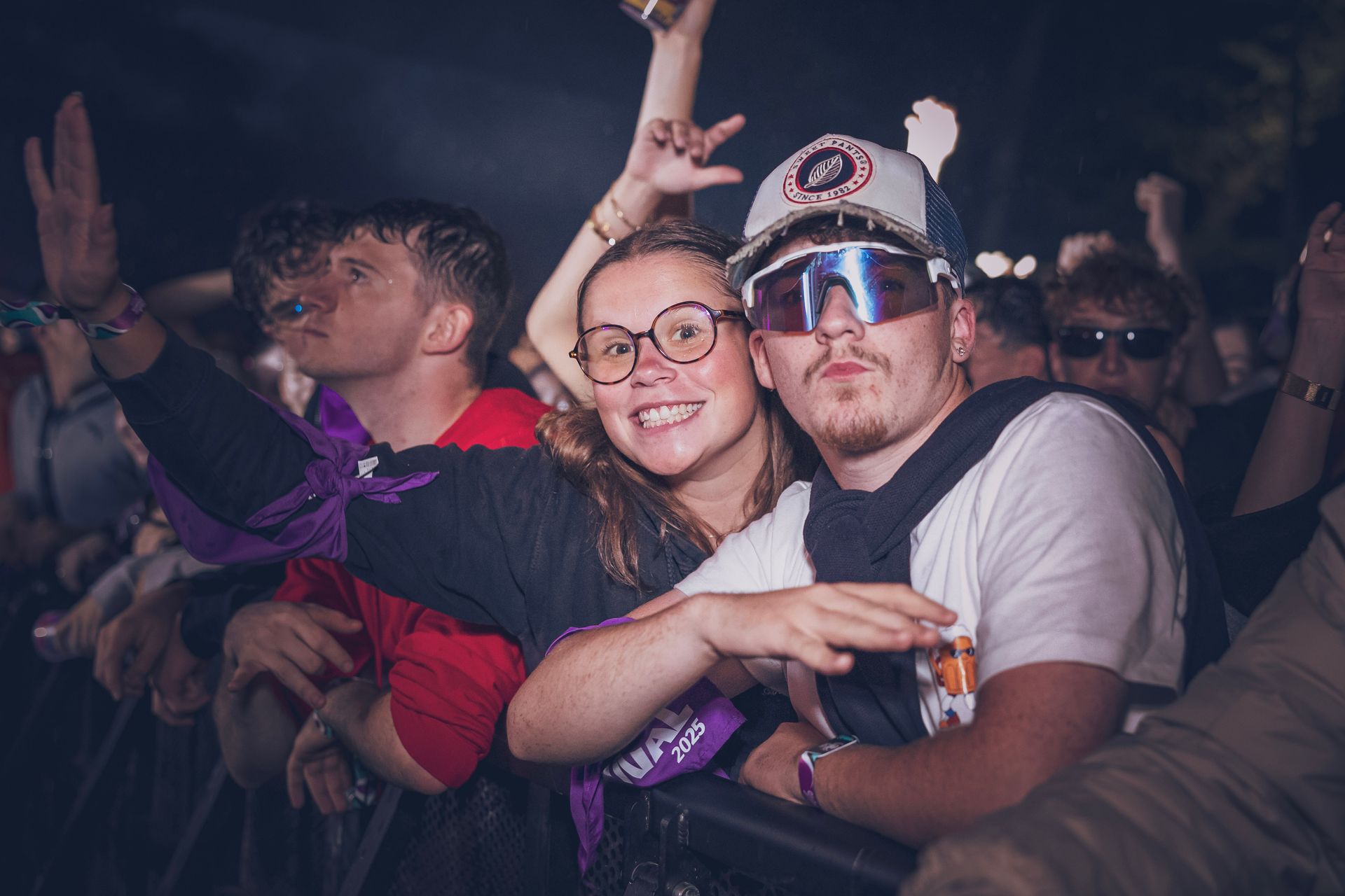 Un groupe de personnes à un concert, souriant et faisant des signes de la main, avec une personne au premier plan portant des lunettes de soleil réfléchissantes.