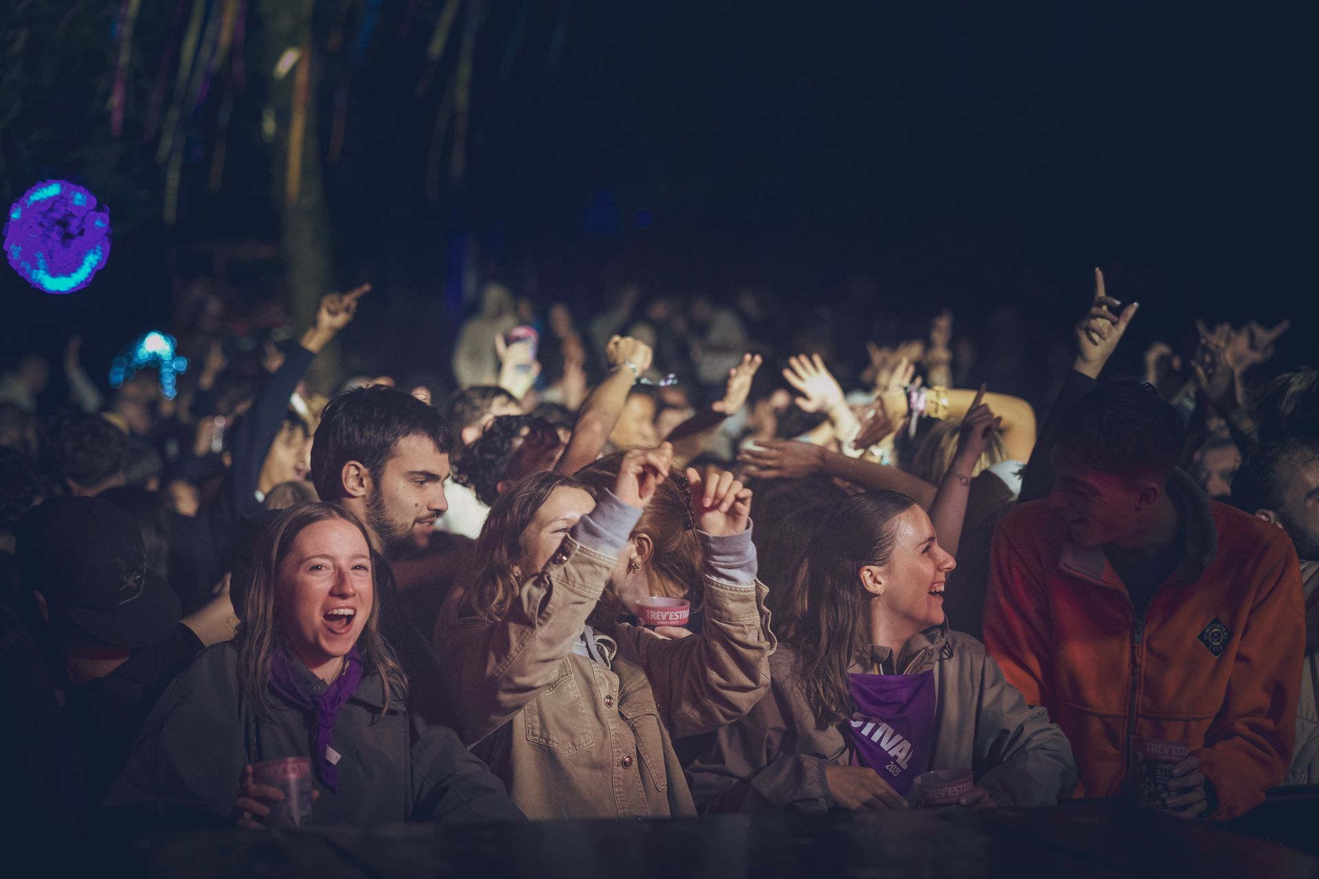 Une foule de personnes à un concert en plein air, de nuit, acclamant la foule les bras levés, éclairée par des projecteurs et des touches de violet.