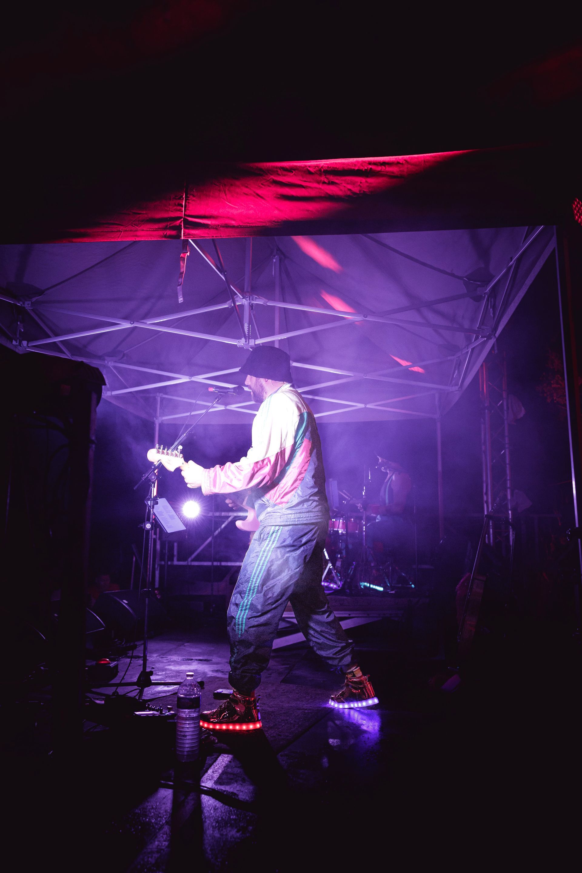 Un artiste se tient sous un dais illuminé de violet sur scène, jouant de la guitare électrique dans une ambiance tamisée et dramatique.