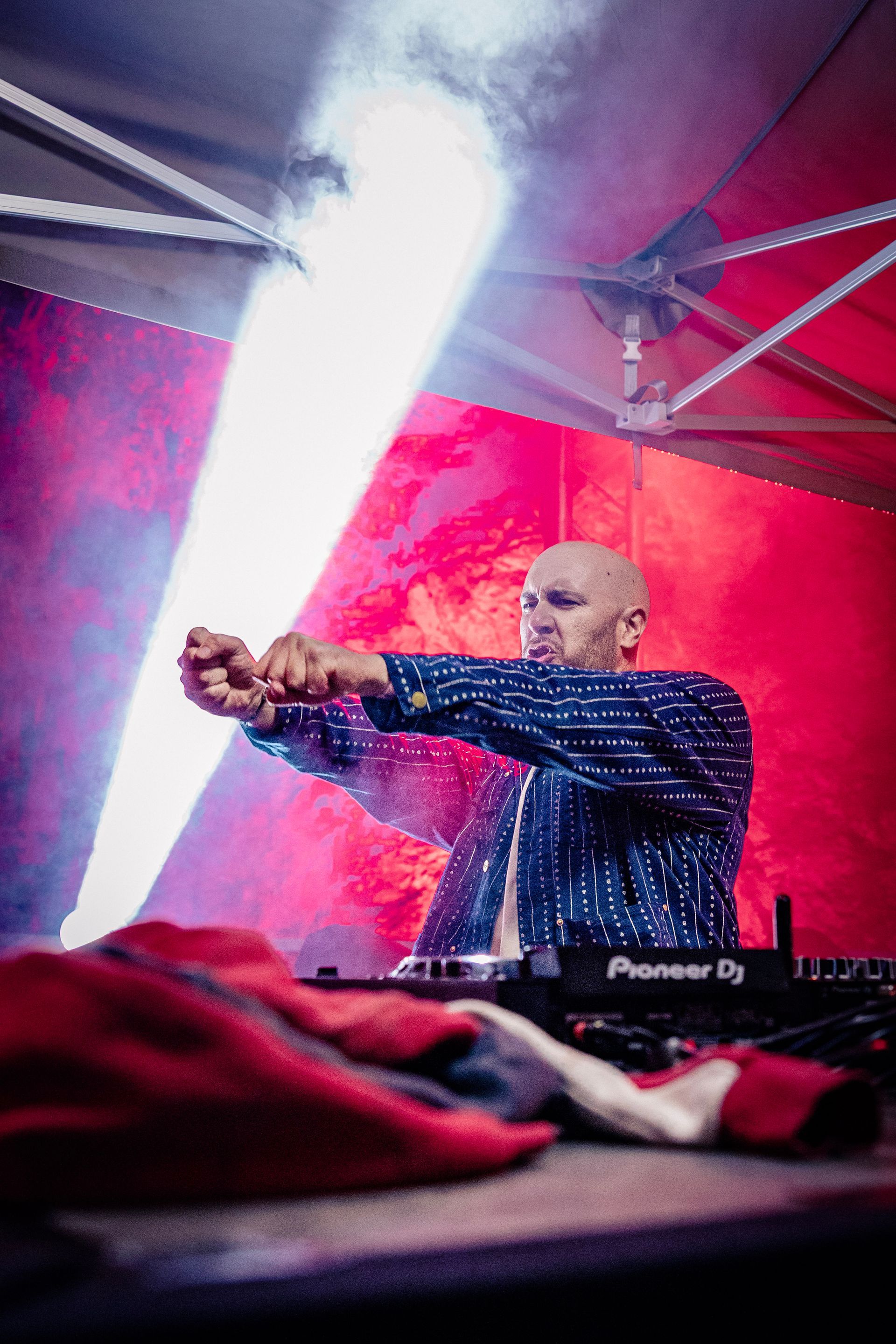 Un DJ chauve, vêtu d'une chemise à motifs, mixe derrière une console, éclairé par un projecteur de scène puissant sur un fond rouge éclatant.