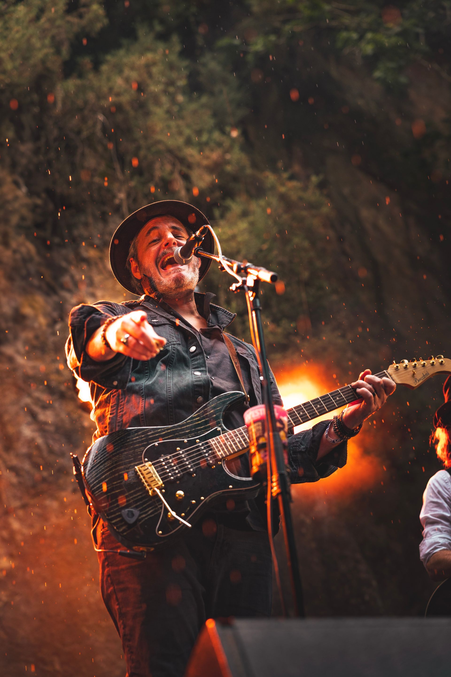 Un musicien coiffé d'un chapeau se produit sur scène, jouant de la guitare électrique et pointant du doigt le public dans une lumière chaude.