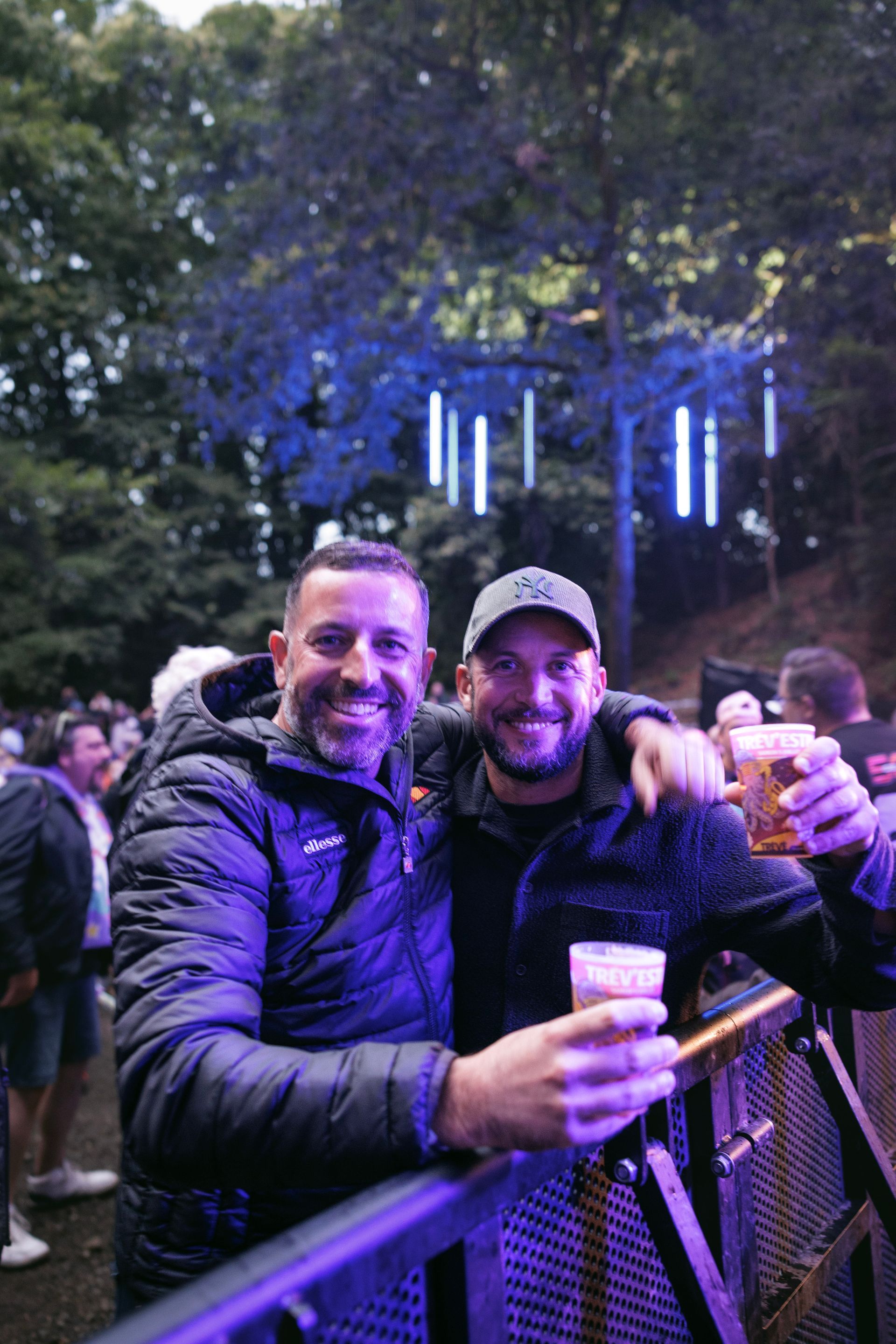 Deux hommes souriants tiennent des verres lors d'un événement en plein air, debout derrière une balustrade métallique sous des arbres illuminés par des guirlandes lumineuses bleues.