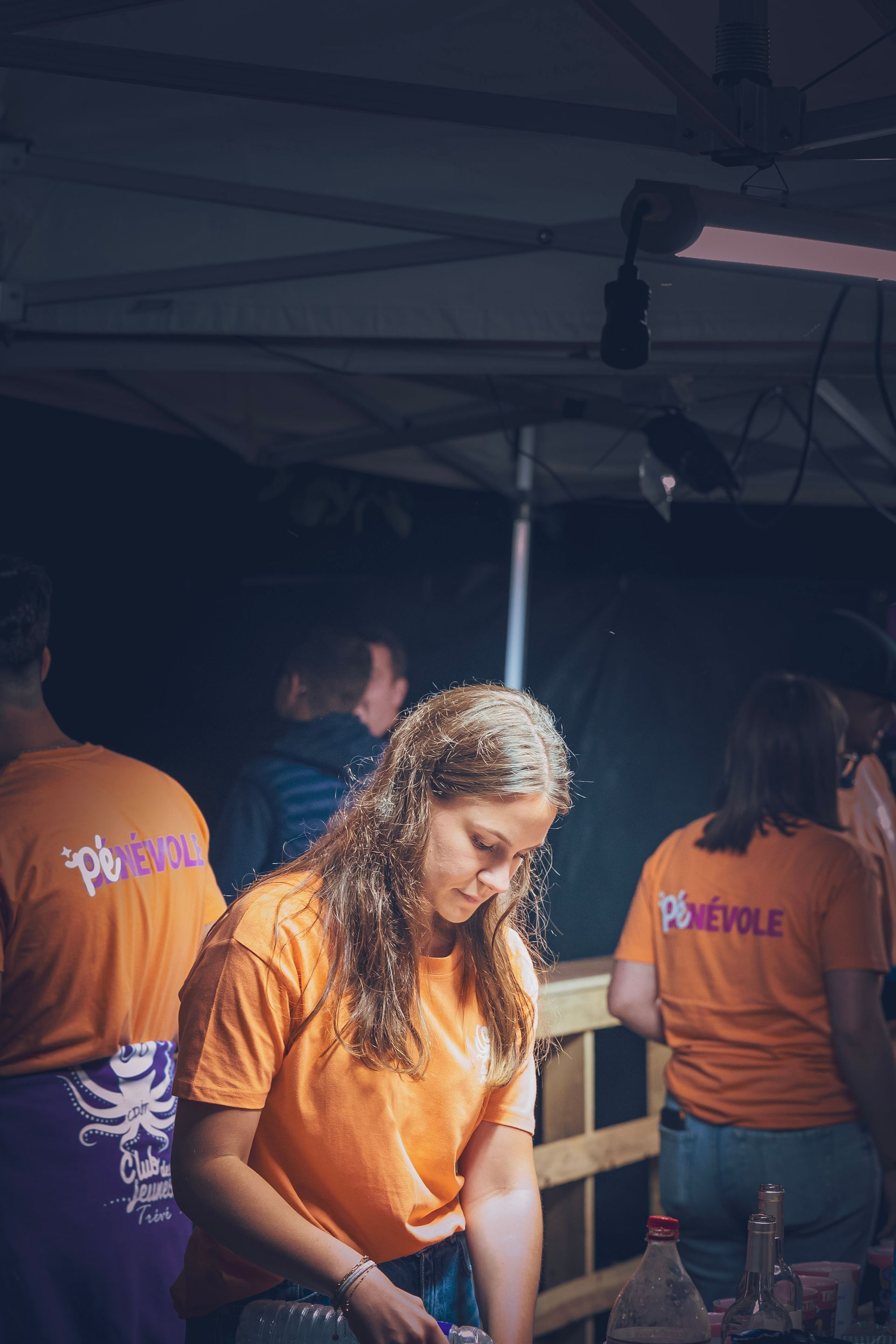 Trois bénévoles vêtus de t-shirts orange travaillent de nuit sous une tente éclairée lors d'un événement en plein air.