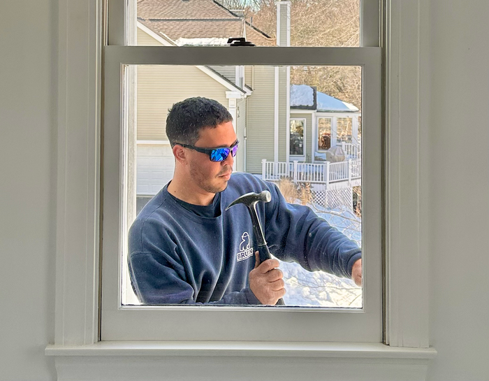 Person in sunglasses using a hammer outside a window, seen through the glass.