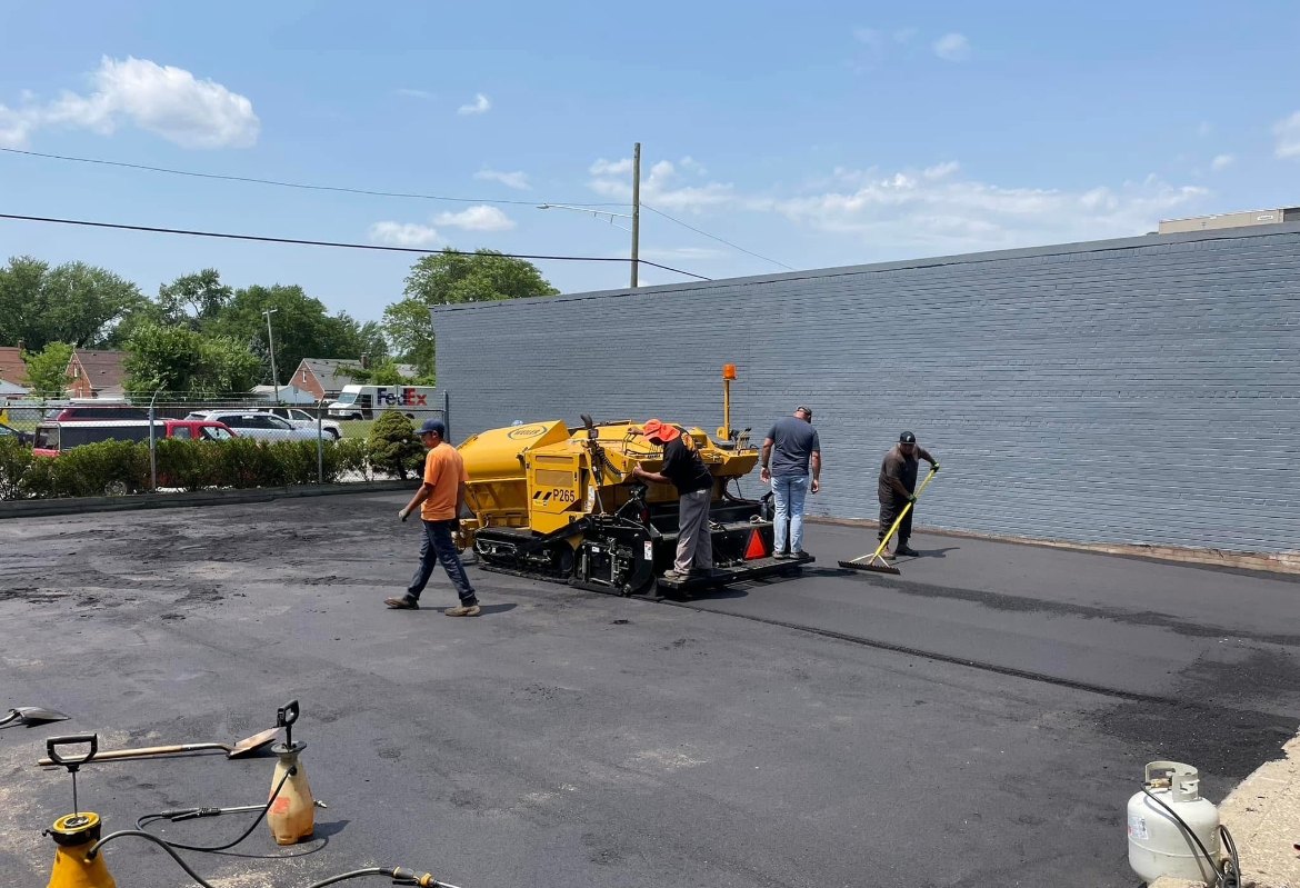 Workers around a yellow road roller on freshly paved asphalt beside a sloped retaining wall