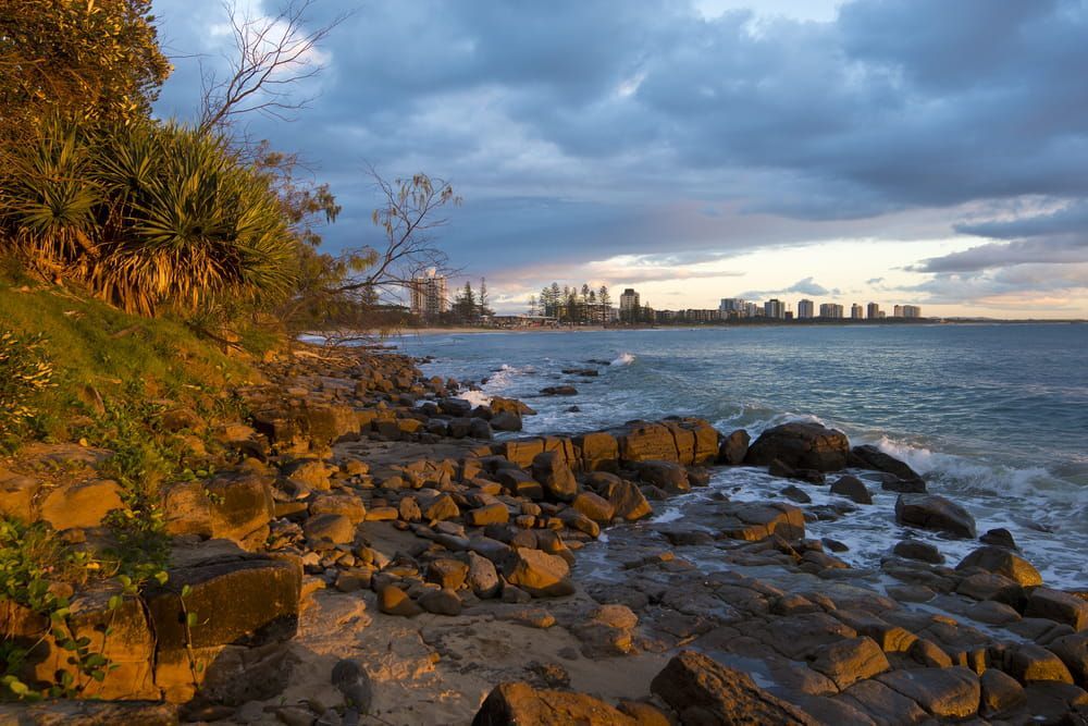 A Rocky Beach With a City in the Background and a Cloudy Sky — Pools By Design in Maroochydore, QLD
