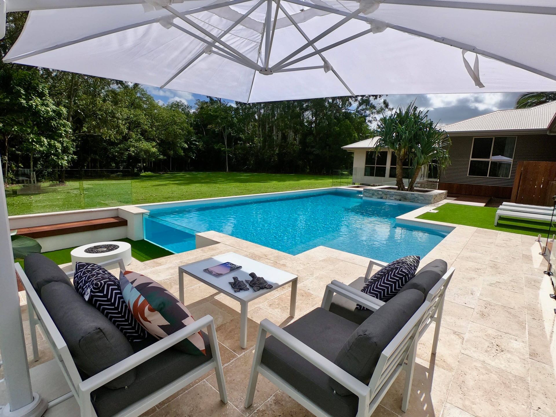 A Large Swimming Pool is Surrounded by Patio Furniture Under an Umbrella — Pools By Design in Warana, QLD