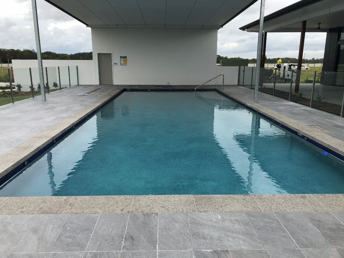 A Large Swimming Pool With a White Building in the Background — Pools By Design in Warana, QLD