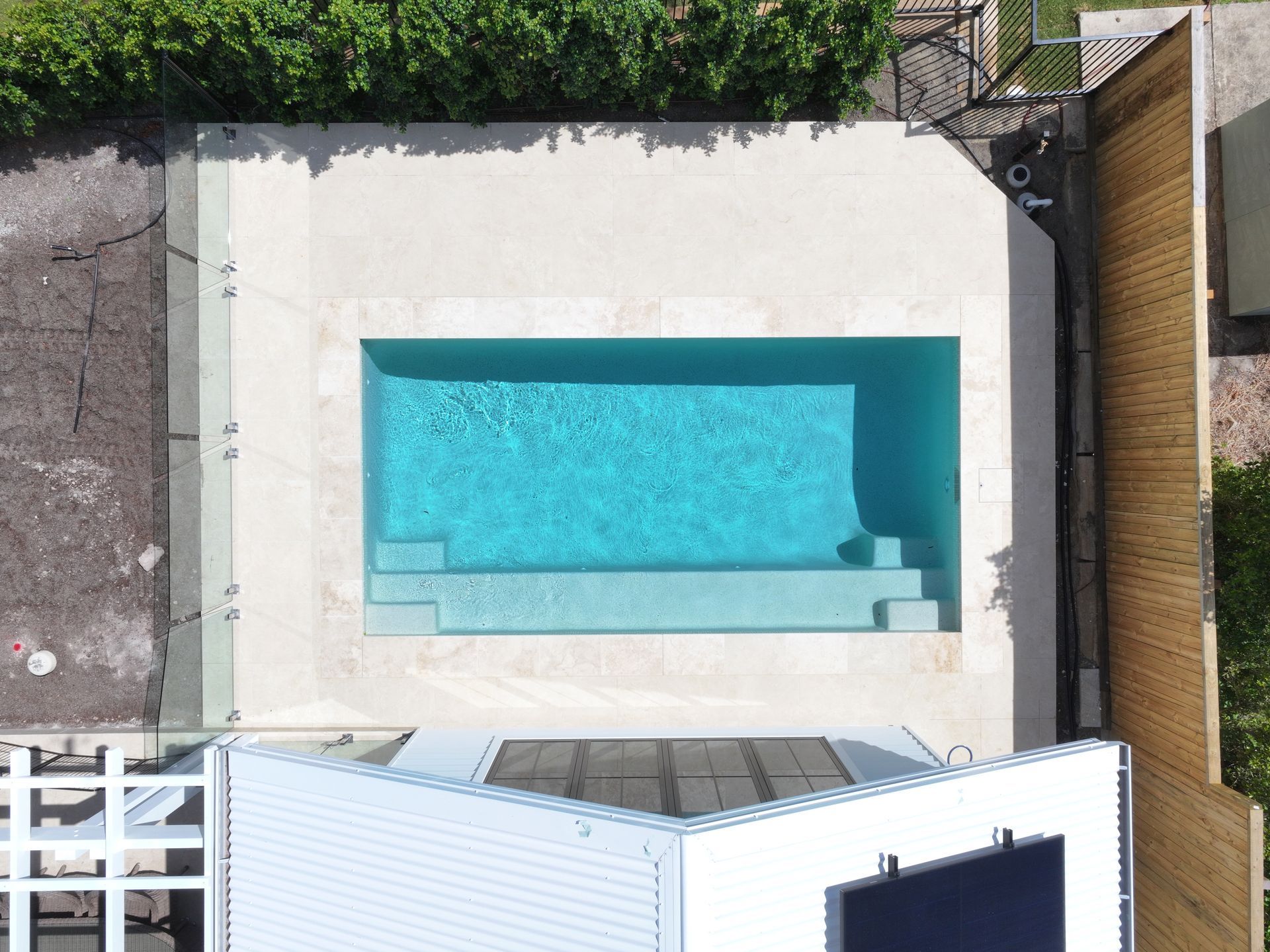 Overhead view of a rectangular turquoise pool surrounded by light-colored stone, next to a white building and wooden fence — Pools By Design in Warana, QLD