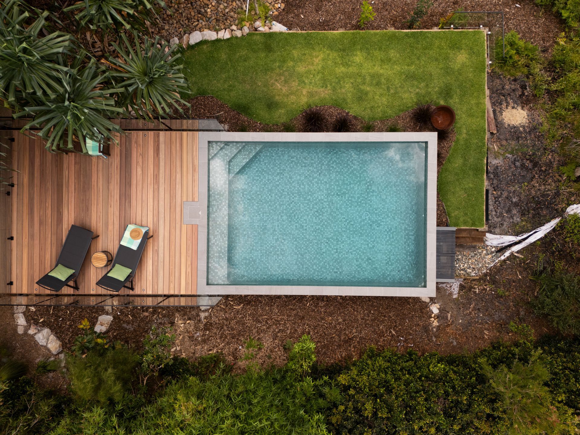 Overhead view of a rectangular pool surrounded by a wooden deck, grass, and trees. Two lounge chairs are on the deck — Pools By Design in Warana, QLD