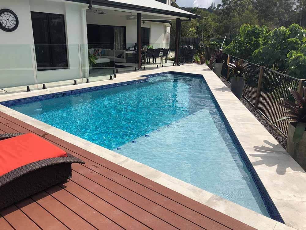 A Large Swimming Pool is Surrounded by a Wooden Deck and a House — Pools By Design in Warana, QLD