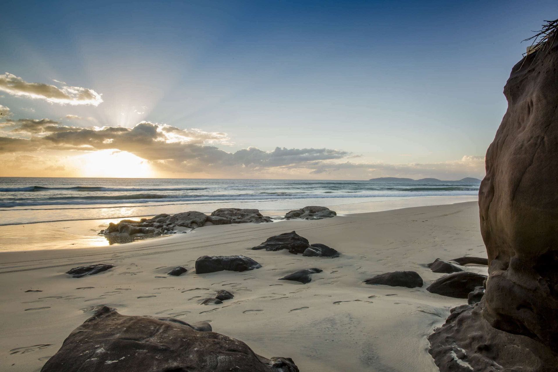 The Sun is Setting Over the Ocean on a Beach With Rocks in the Foreground — Pools By Design in Gympie, QLD