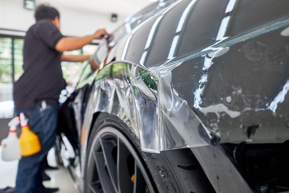 Person Applying Clear Protective Film to a Black Car's Fender, Indoors — Airlie Beach Smash Repairs in Cannonvale, QLD