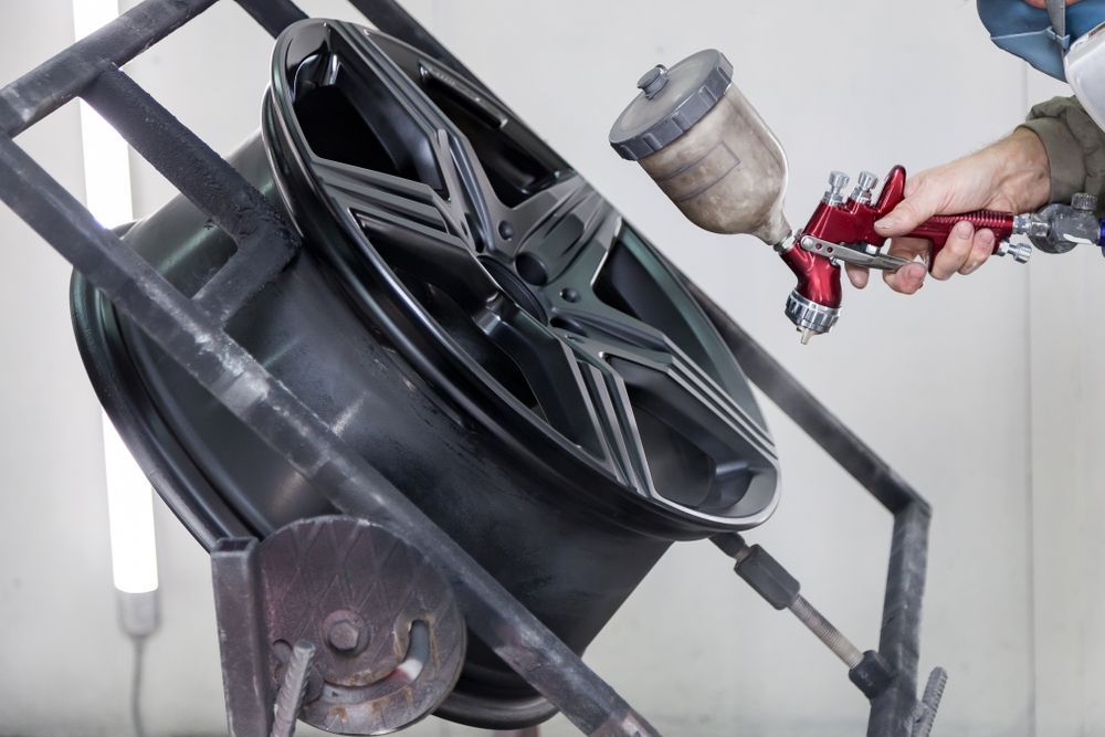 Person Spray Painting a Black Car Wheel in a Workshop, Using a Red Spray Gun — Airlie Beach Smash Repairs in Proserpine, QLD
