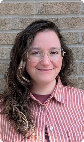 Woman with dark hair smiles, leaning against a brick wall, wearing a white button-down shirt.