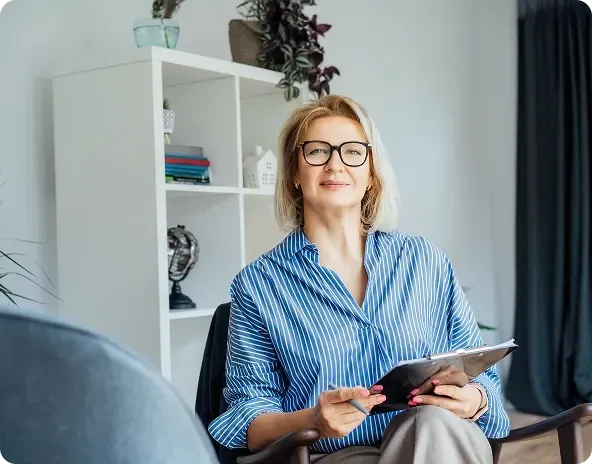A smiling therapist with blonde hair and glasses sits in a chair, holding a clipboard, in her office.