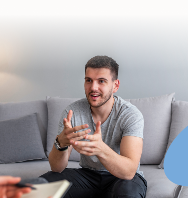 Man seated on a gray sofa, gesturing with hands while speaking to someone, likely in a therapy session.
