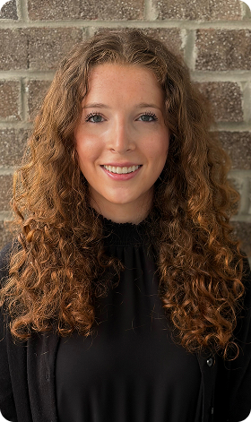 Woman wearing graduation sash, smiling outdoors, with spring blossoms in background.