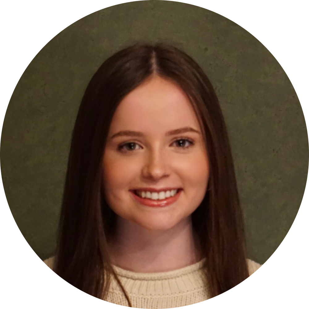 Woman with long brown hair smiles at the camera, wearing a cream sweater against a green background.