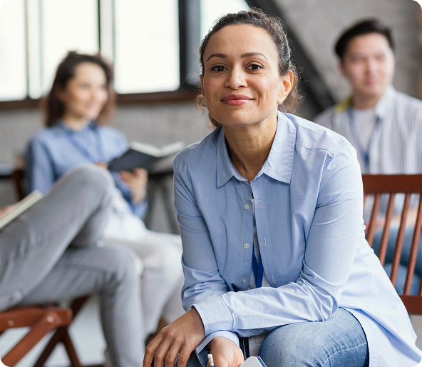 Woman in a blue shirt smiles, seated in a chair. Two other people are in the background, also seated.