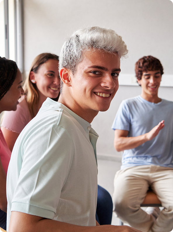 A smiling young man with light-colored hair looks over his shoulder in a group setting, surrounded by other people.
